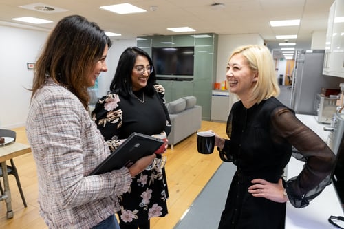 Three women shown in the Edenred office's kitchen area; one woman is holding a coffee mug, another is holding a laptop and the third is laughing. They are all shown having a discussion