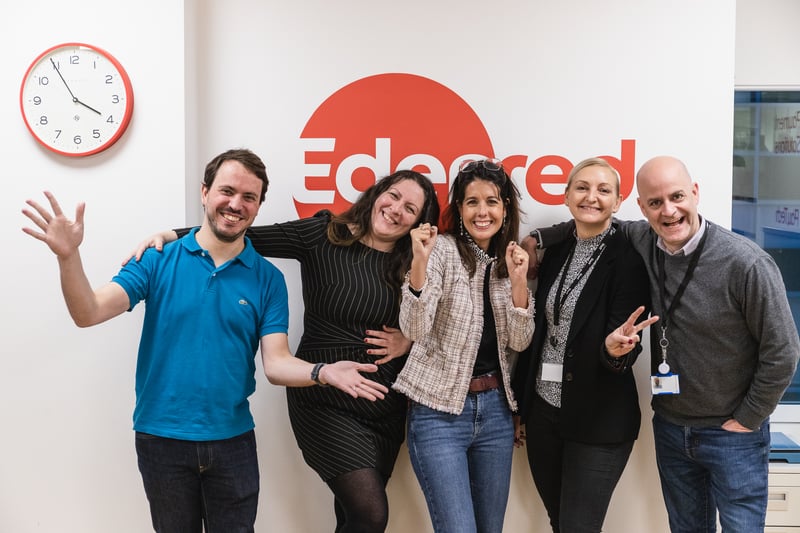 A team group photo that shows five employees standing next to each other with their hands raised in the air. On the wall behind them, the wall reads 'Edenred' in red text besides a small red clock and window.