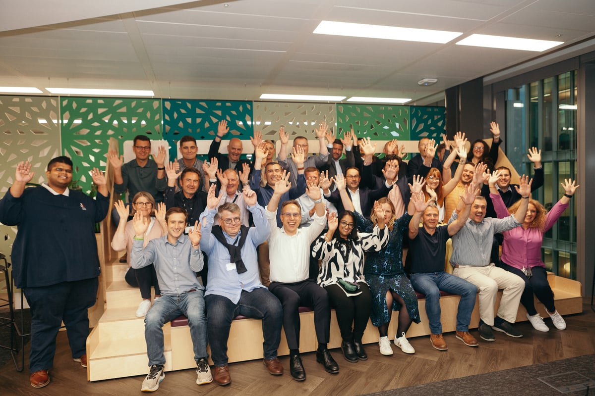 A team photo with dozens of employees shown, all sitting in a communal area with their arms raised during an event. The office has white, light green, and dark green dividers in the background and a glass wall on one side.