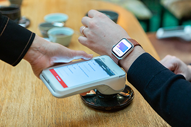 An image showing one person holding a card reader device to another persons digital watch; on the watch a digital payment card is shown