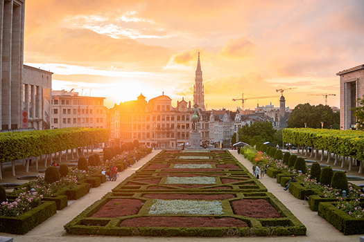 a cityscape shown as sunset time that includes large buildings, a garden, and some people walking through the environment