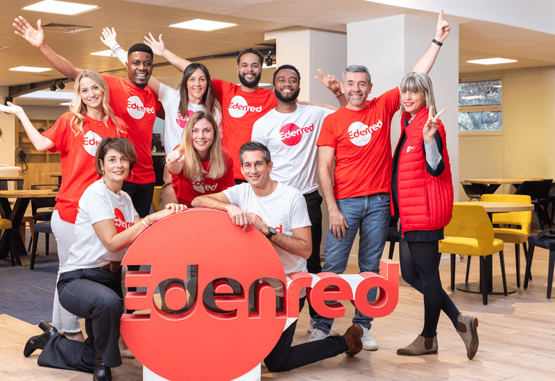 Edenred group photo showing several employees standing near the Edenred Logo as a standup prop; the prop is Red and the employees are wearing white and red tshirts and include both men and women