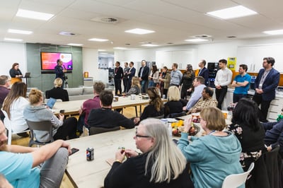 a wide shot of an Edenred office with several people watching a presentation being given; some are standing by a kitchen area against a wall and others are sitting in chairs by tables, all of them are facing the presenter at the head of the room